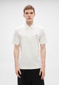 Λευκό πόλο πουκάμισο με κοντά μανίκια, τρεις κουμπότρυπες και μικρό λογότυπο στο αριστερό στήθος. Λείο ύφασμα και κλασική γραμμή.