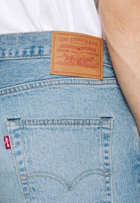 Jeans de mezclilla azul claro con el bolsillo trasero que tiene la etiqueta roja de Levi's y un parche de cuero en la cinturilla, persona vistiendo una camisa blanca.