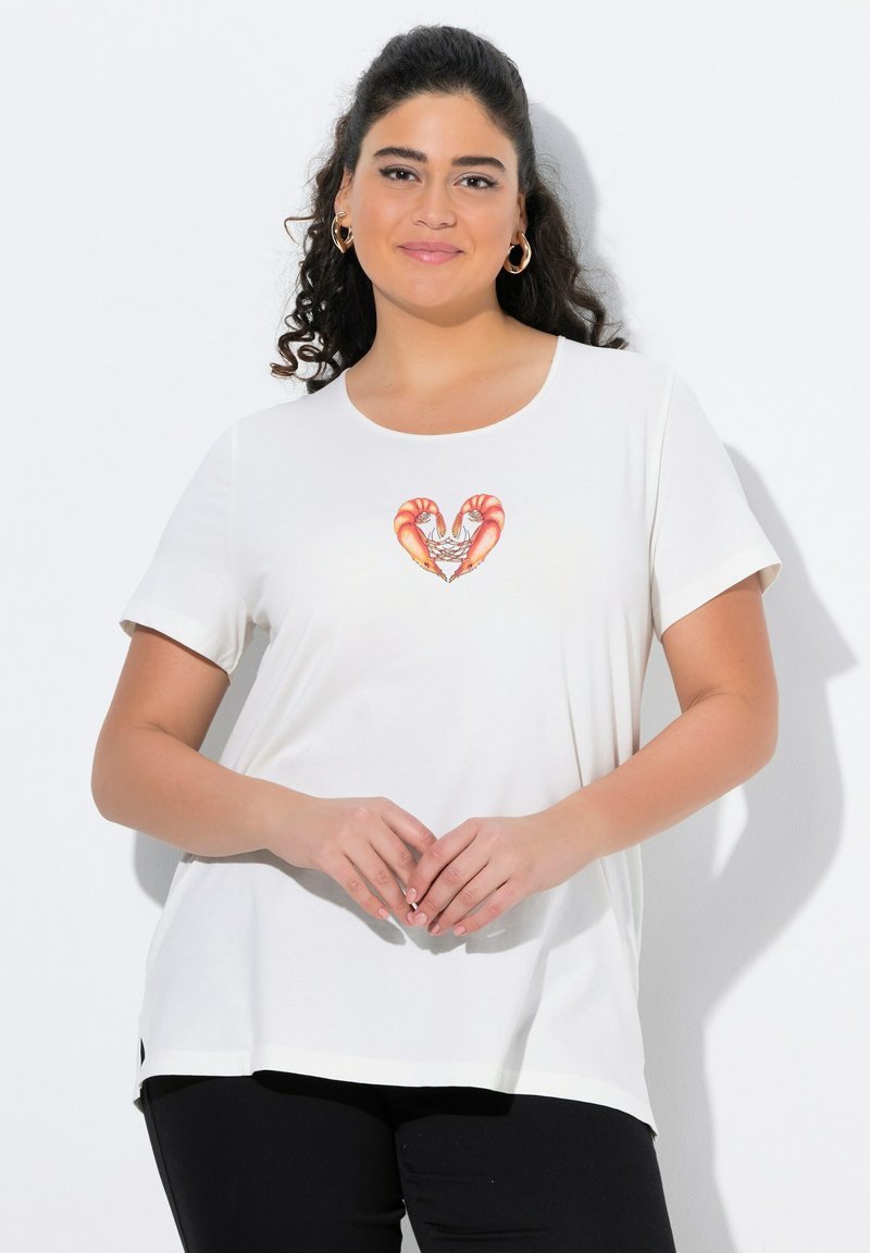 Usměvavá žena s dlouhými kudrnatými vlasy, oblékající zlaté kruhové náušnice, bílý tričko se designem srdce s krevety a černé kalhoty, stojící s rukama sklopenýma.