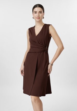 Femme debout portant une robe portefeuille sans manches, longueur genou, de couleur marron foncé, avec des boucles d'oreilles créoles dorées et un bracelet, sur un fond blanc uni.