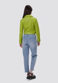 Giacca corta in pelle verde con colletto, abbinata a jeans a vita alta di colore blu chiaro. I jeans presentano un orlo grezzo e tasche posteriori tipiche.