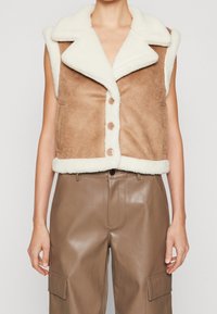 Brun faux suède väst med vit faux shearling-krage, framknappar och sidorfickor, i kombination med glänsande bruna läderbyxor.