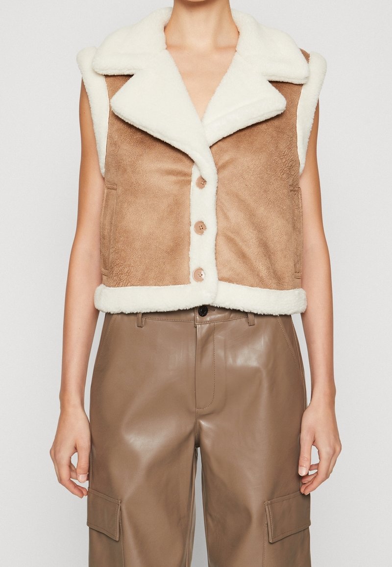 Brun faux suède väst med vit faux shearling-krage, framknappar och sidorfickor, i kombination med glänsande bruna läderbyxor.