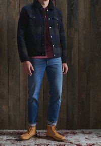 Giacca a quadri blu navy con colletto, t-shirt rossa sotto, jeans blu e stivali gialli. Ambientata su uno sfondo di legno.