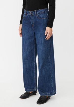 Personne portant un jean bleu pattes d'éléphant taille haute, un haut noir à manches longues et des chaussures noires sans lacets, debout devant un fond blanc.