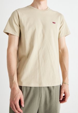 Beige Levi's t-shirt med korte ærmer båret af en person, kombineret med olivengrønne bukser mod en ensfarvet hvid baggrund.