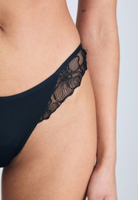 Bikini noir en tissu lisse avec des accents en dentelle transparente présentant des motifs floraux sur les côtés. S'ajuste confortablement autour des hanches.