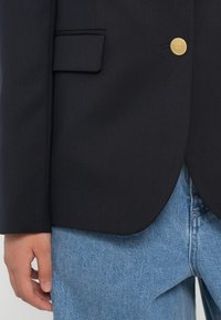 Zwarte blazer met een strakke ontwerp, enkele gouden knoopsluiting en een voorzak, gecombineerd met lichtblauwe spijkerbroek.