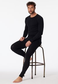 Top negro de manga larga y pantalones ajustados en una suave tela elástica. El modelo está sentado en un taburete de metal, mostrando el diseño minimalista.