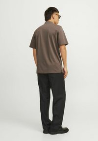 Hnedá polokošeľa s krátkym rukávom, bavlnený materiál, voľný strih, postranné rozparky, kombinovaná s čiernymi nohavicami a čiernymi topánkami. Pohľad diváka zozadu.