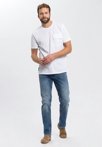 Witte t-shirt met korte mouwen en een borstzak, gecombineerd met lichtblauwe spijkerbroek en bruine suède laarzen, op een neutrale achtergrond.