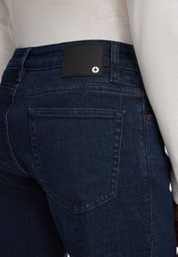 Gros plan sur une poche et une ceinture de jean en denim bleu foncé avec une patch noire présentant un détail de croix blanche, porté avec un dessus blanc.