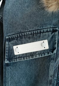 Chaqueta de mezclilla con un lavado azul oscuro, que presenta un gran bolsillo en el pecho, una etiqueta blanca con texto en relieve y detalles de costura visibles.