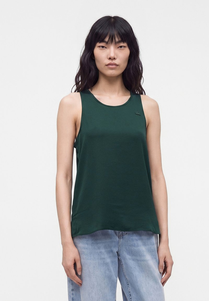 Femme aux longs cheveux foncés portant un haut sans manches vert foncé et un jean bleu clair sur un fond clair uni.