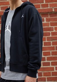 Person som har på sig en svart zip-up hoodie över en grå t-shirt med en vit sportlogotyp, stående mot en bakgrund av röd tegelvägg.