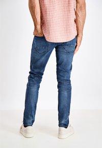 Jeans ajustados de mezclilla azul, con una cintura de altura media, cinco bolsillos y un sutil desgastado. Combina con una camisa de cuadros rosa y zapatillas blancas.