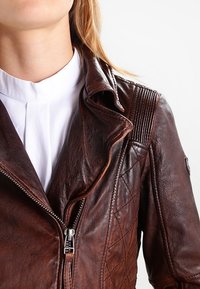 Giacca in pelle marrone con colletto a punte, chiusura con zip e dettagli a coste sulle spalle. Presenta un motivo trapuntato sul corpo.