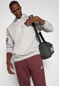 Homem usando um fato de treino cinza com capuz e calças bordô com logótipos, ajustando a alça de uma mochila preta da Adidas em fundo simples.