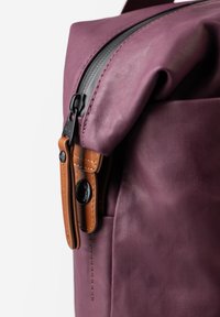 Zaino in tessuto viola con superficie texurizzata, caratterizzato da una cerniera nera e un accentuato tirazip in pelle. Dettagli cuciti visibili.