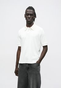 Mladý muž s tmavou pletí a dredy, oblečený v bílé polokošili s krátkými rukávy a volných černých džínách, stojí s jednou rukou v kapse.