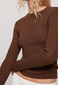 En person med vågigt hår har på sig en figurnära brun ribbad tröja och håller försiktigt om sin midja, vilket kompletteras av en enfärgad bakgrund.