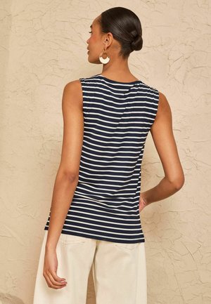 Vrouw met haar in een knot, dragend gouden en witte oorringen in hoepelvorm, een mouwloze marineblauwe en wit gestreepte top en lichtbeige broek tegen een gestructureerde muur.