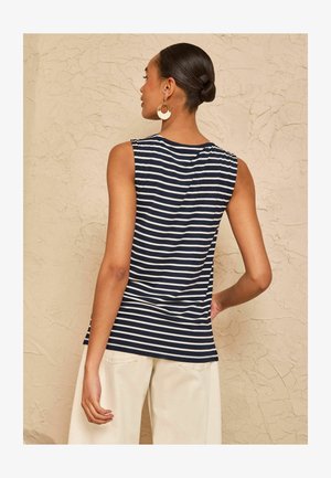 Vrouw met haar in een knot, dragend gouden en witte oorringen in hoepelvorm, een mouwloze marineblauwe en wit gestreepte top en lichtbeige broek tegen een gestructureerde muur.