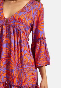 Paarse blouse met een V-hals, voorzien van een levendig oranje bloemenpatroon, klokmouwen en geraffineerde details bij de buste en zoom.