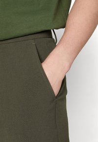 As calças verde-azeitona apresentam um bolso lateral, uma textura suave e um detalhe de costura discreto. O material parece elástico e durável.