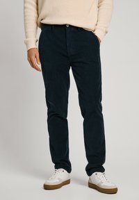 Person wearing dark blue corduroy pants, beige sweater, and white sneakers with gum soles standing against a light background.