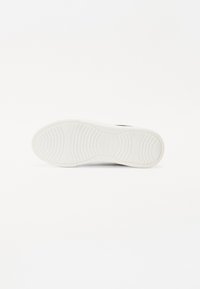 Semelle de sneaker en caoutchouc blanc avec des rainures texturées et des grips en motif ; présente un logo et un détail de marque près du talon.