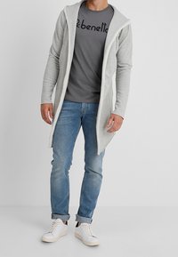 Hellgrauer langer Cardigan mit Kapuze, getragen über einem dunkelgrauen T-Shirt mit schwarzem Text, kombiniert mit blauen Jeans und weißen Turnschuhen.