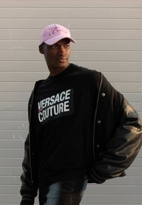 Svart bomberjakke med lærermmer, svart t-skjorte med "VERSACE JEANS COUTURE"-logo, rosa caps med skråskrift "Couture"-tekst, blå jeans.