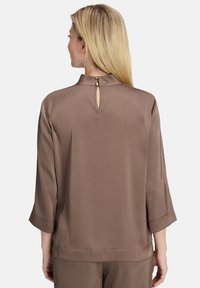 Blusa leggera marrone con colletto alto, vestibilità ampia, maniche a trequarti e apertura a occhio di chiave sul retro, decorata con bottoni.