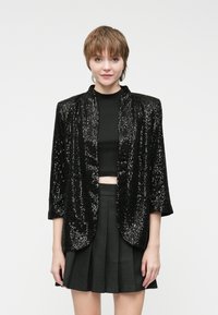 Crna jakna s pailetnim detaljima i bez ovratnika, tri četvrtine rukava i slobodnim krojem, nosi se preko crnog crop topa i sivih suknji.