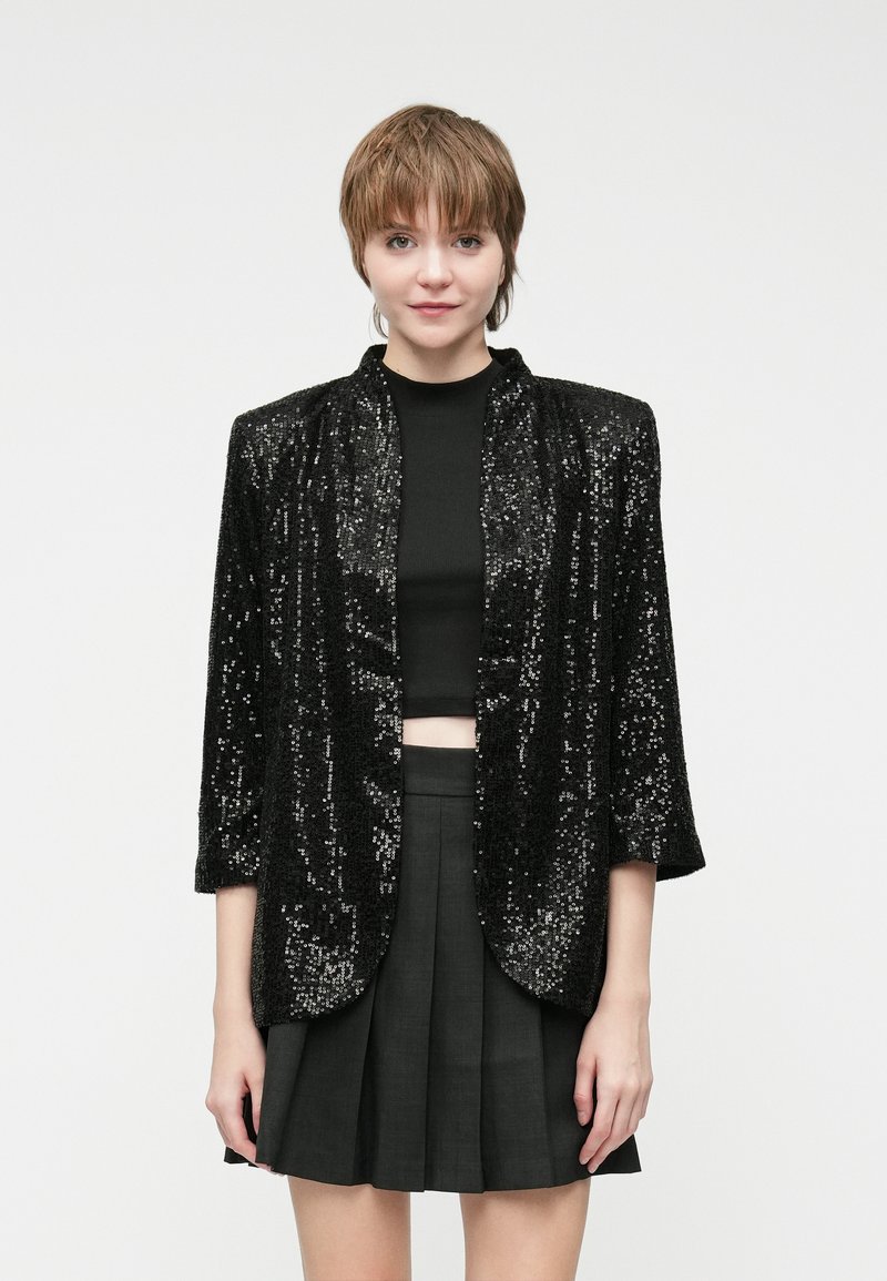 Crna jakna s pailetnim detaljima i bez ovratnika, tri četvrtine rukava i slobodnim krojem, nosi se preko crnog crop topa i sivih suknji.