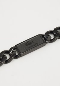 Pulsera de cadena negra con un cierre rectangular texturizado que presenta el logo de Lacoste. Enlaces lisos y acabado brillante en toda la pulsera.