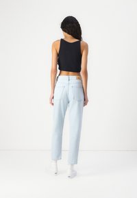 Femme debout, dos tourné, portant un crop top noir sans manches, un jean taille haute bleu clair, des chaussettes blanches et des baskets blanches.