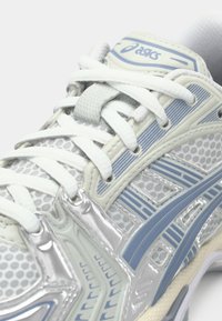 Weißer Sportschuh mit Mesh und glänzenden silbernen Akzenten. Verfügt über blaue Designlinien, strukturierte Schnürsenkel und eine gepolsterte, moderne Silhouette.