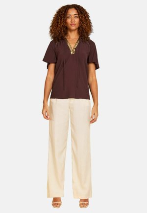 Kobieta z kręconymi włosami, ubrana w brązową bluzkę z krótkim rękawem z złotą lamówką i kremowe spodnie o szerokich nogawkach, stojąca na białym tle.