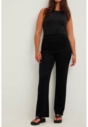 Mujer que lleva una camiseta negra sin mangas, pantalones negros de talle alto y sandalias negras de plataforma sobre un fondo liso y claro.