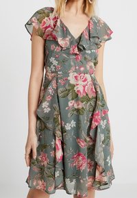 Kobieta w zielonej sukience w kwiaty z różowymi i białymi kwiatami, marszczonym dekoltem w kształcie litery V oraz falbanistymi rękawami.