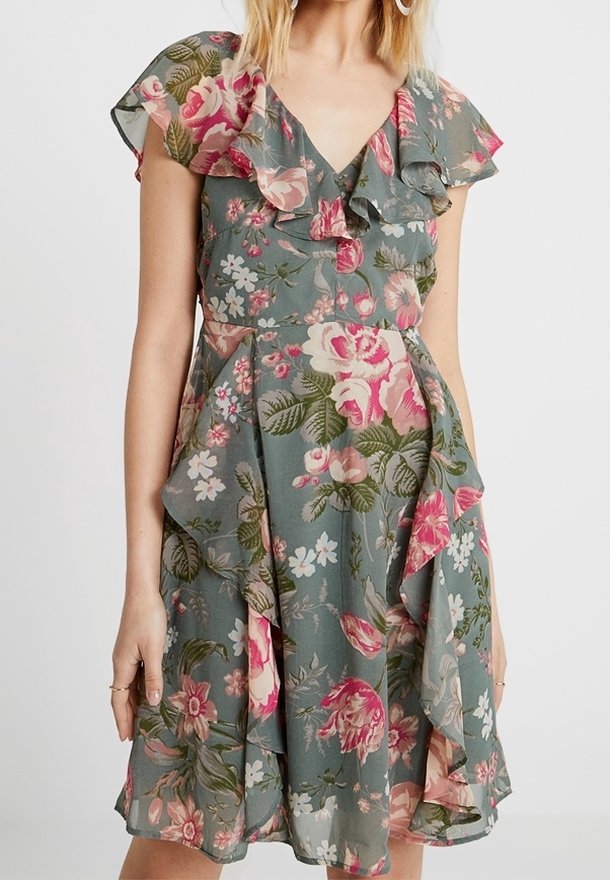 Kobieta w zielonej sukience w kwiaty z różowymi i białymi kwiatami, marszczonym dekoltem w kształcie litery V oraz falbanistymi rękawami.