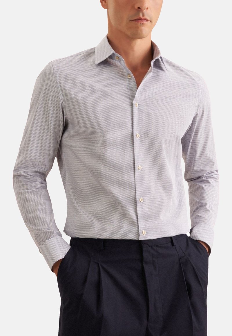 Camicia a maniche lunghe di un azzurro chiaro con un sottile motivo geometrico, colletto button-down e bottoni dorati. Indossata con pantaloni pieghettati scuri.