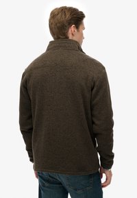 Bruine fleecejas met een hoge kraag, textuur op het oppervlak en contrasterende stiksels. Heeft een losse pasvorm en lange mouwen.