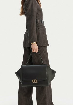 Mujer vestida con un traje de rayas oscuras sostiene un bolso de cuero negro con un logo dorado GR, de pie frente a un fondo blanco liso.