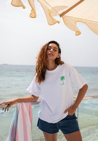 Mujer con camiseta blanca con diseño de palmera y pantalones cortos de mezclilla apoyada en la barandilla junto a la playa, con una toalla a rayas y una sombrilla arriba.