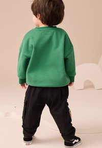 Zöld pulóver esett vállakkal, fekete cargo nadrággal, oldal zsebekkel. Fekete tornacipő fehér talppal és zöld részletekkel.