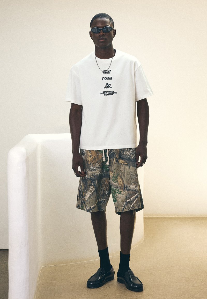Man som bär svarta solglasögon, vit t-shirt med grafik, shorts med kamouflagemönster, svarta strumpor och svarta loafers, som står inomhus.