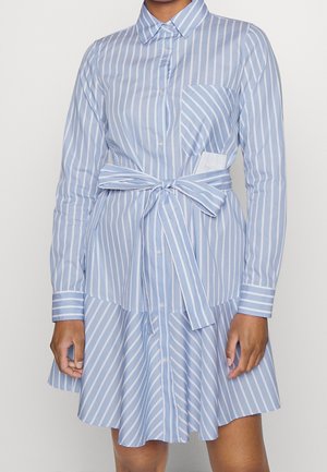 Frau trägt ein hellblau-weiß gestreiftes Hemdblusenkleid mit vertikalen Streifen, einem gebundenen Taillengürtel und einer Brusttasche.